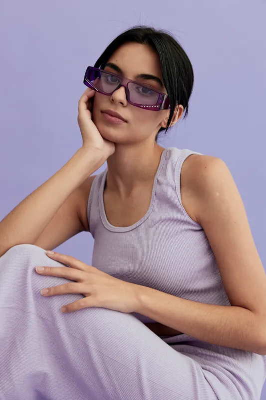 stylish woman with purple sunglasses wearing lilac tank top and skirt sitting against pastel purple background, showing relaxed pose and modern fashion