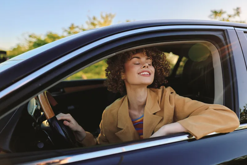 Young woman with curly hair sits in a car, smiling with eyes closed as warm sunlight fills the cabin, creating a peaceful, carefree moment with soft reflections on the seats.