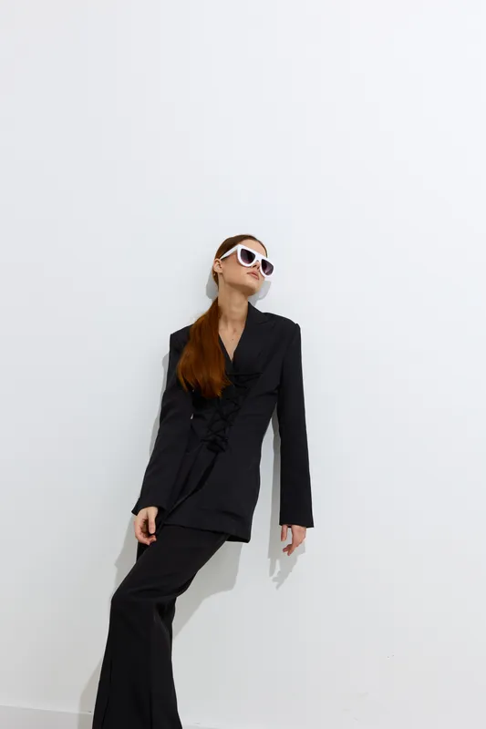 Woman in black suit posing confidently against white wall, wearing stylish sunglasses with sleek red hair and minimalistic background, showcasing modern fashion elegance.