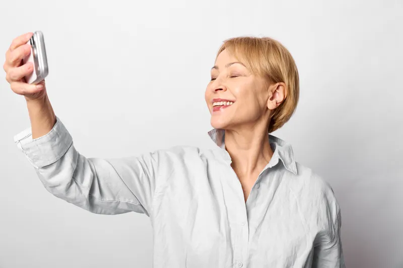Smiling woman taking a selfie with a smartphone, close portrait in a studio with white background, relaxed happy mood, casual shirt, soft studio lighting, daylight
