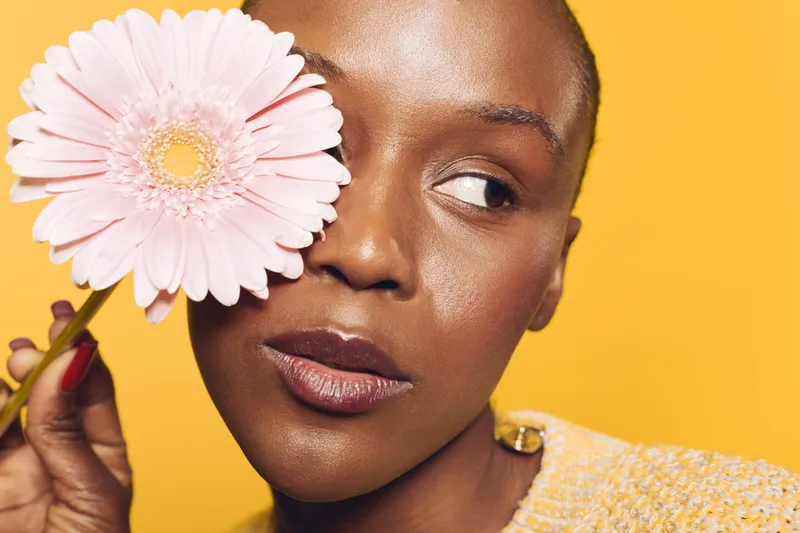 Close-up of dark skinned African woman holding pink flower over eye on yellow background. Concept of beauty, skincare, natural, wellness, and floral aesthetics in studio lighting.