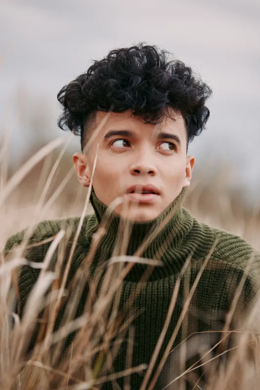 Close natural portrait of a young man framed in tall beach grass, looking thoughtfully aside, capturing an authentic intimate moment with credible emotional expression and soft natural light.
