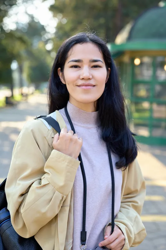 Woman with backpack in an outdoor park setting, wearing a casual jacket and sweater, smiling at the camera, embodying student lifestyle, learning, growth, and everyday exploration on a sunny day