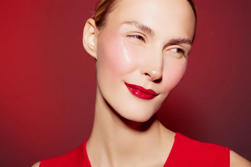 Close-up of a smiling woman with red lipstick and smooth skin wearing a red top against a red background. Concept of beauty, makeup, skincare, and confident expression in studio lighting.