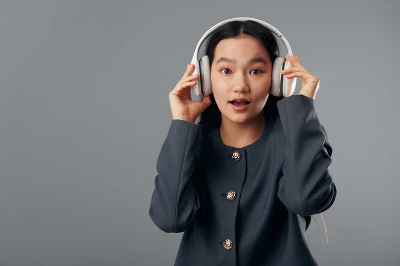 Young woman wearing large headphones with surprised expression listening to music or sound in studio with gray background. Casual clothing and long hair with focused emotion.
