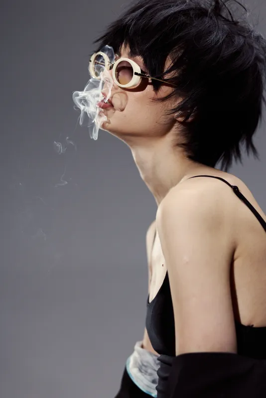 Young woman exhaling smoke wearing round glasses and black top, stylish and expressive portrait with dark background.