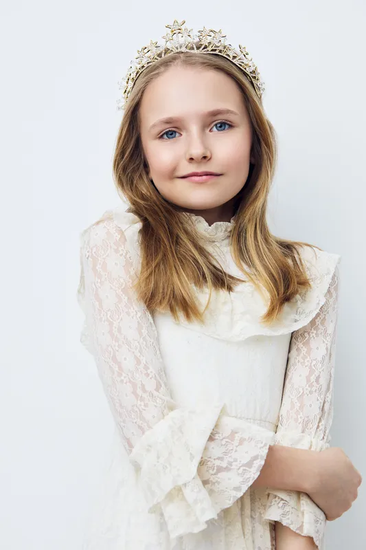 Young girl wearing a lace dress and crown, smiling gently against a light background, evoking feelings of innocence and joy, perfect for seasonal themes