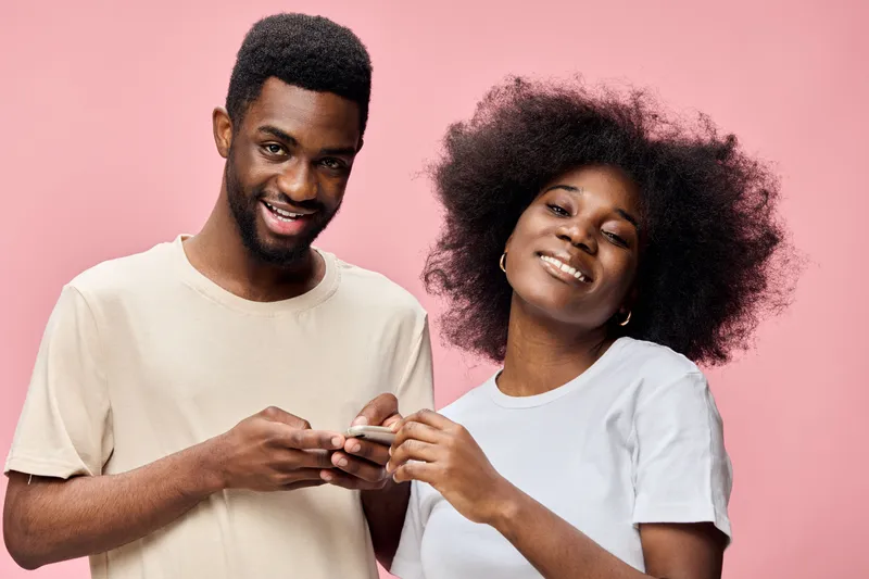 Happy young couple with smartphones, showcasing connection and affection against a pastel pink background, emphasizing joy and modern lifestyle