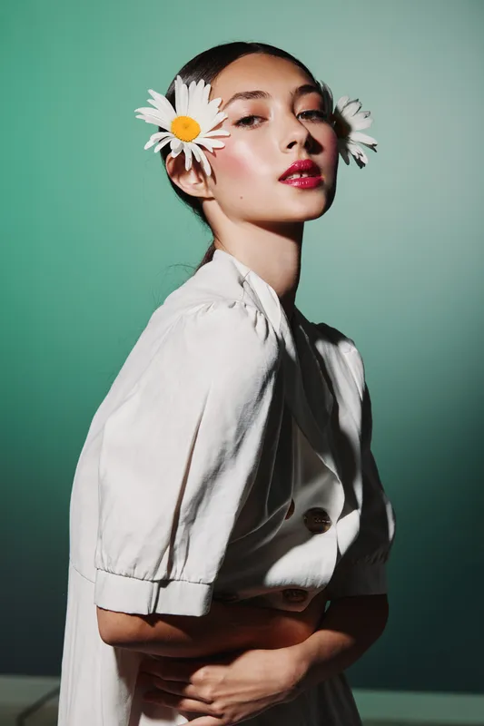 Elegant young woman with daisies in hair poses against a green background. Her bright makeup and white outfit emphasize beauty, creativity, and fashion style in a studio setting.