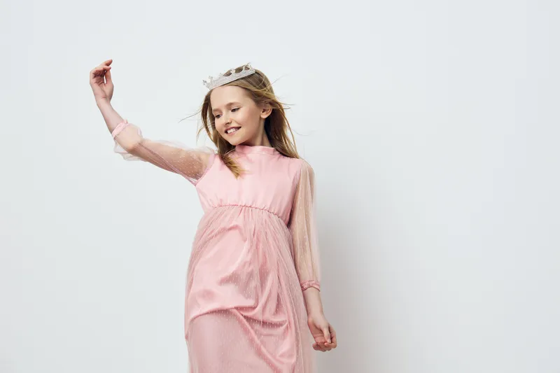 Cheerful girl in pink dress with crown, enjoying playful moment in studio, symbolizing innocence and joy in childhood, perfect for fashion or children s themes
