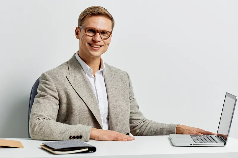 Professional smiling man in beige suit and white shirt working at modern office with laptop, glasses, and notepad on desk. Business, technology, and success concept.