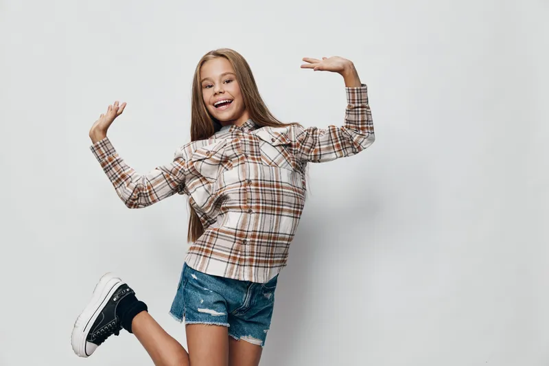 Girl, child, smiling and posing in casual clothes with plaid shirt and denim shorts on studio background for happiness and youth activity.