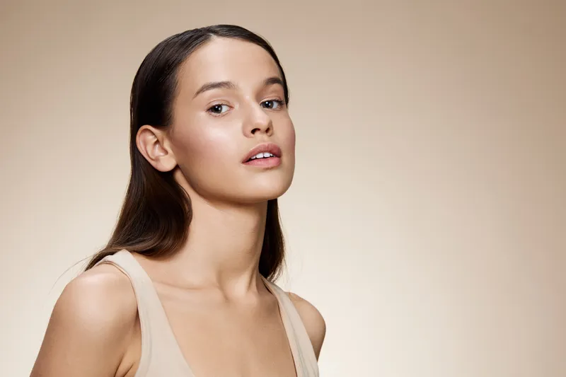 Young woman with long brown hair and natural makeup against a soft beige background, showcasing natural beauty and confidence