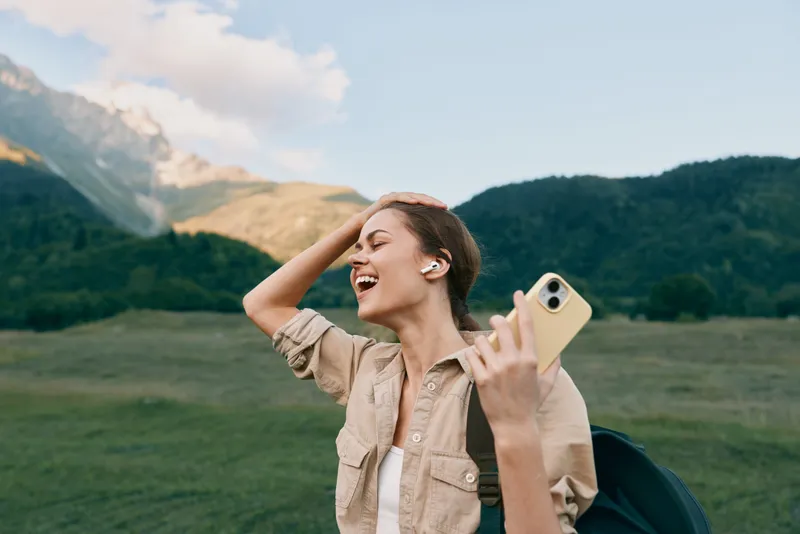 Young woman with backpack smiles while taking a selfie in a green meadow with far mountains, her relaxed posture and sunny mood conveying outdoor adventure and carefree lifestyle