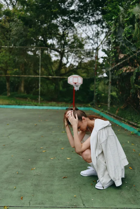 Young female athlete in sportswear, showcasing frustration on a basketball court surrounded by greenery, reflecting determination and emotional resilience
