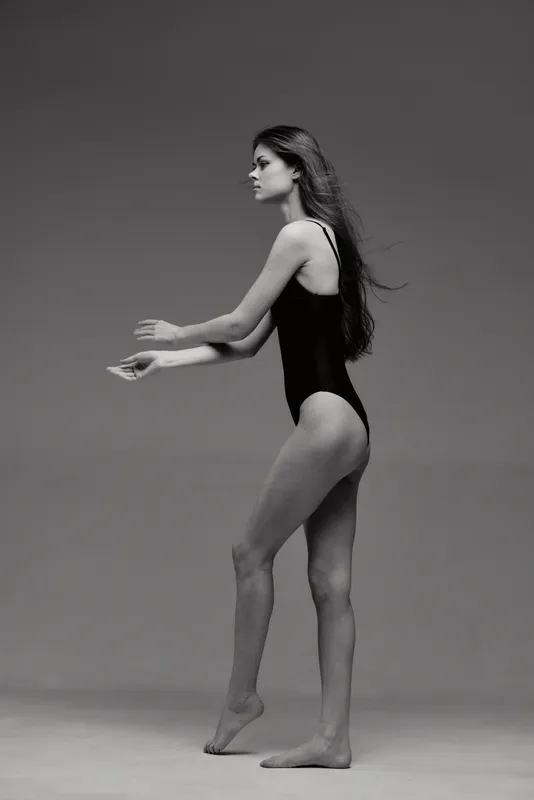 Black and white image of a young woman in a black leotard posing gracefully against a neutral background.