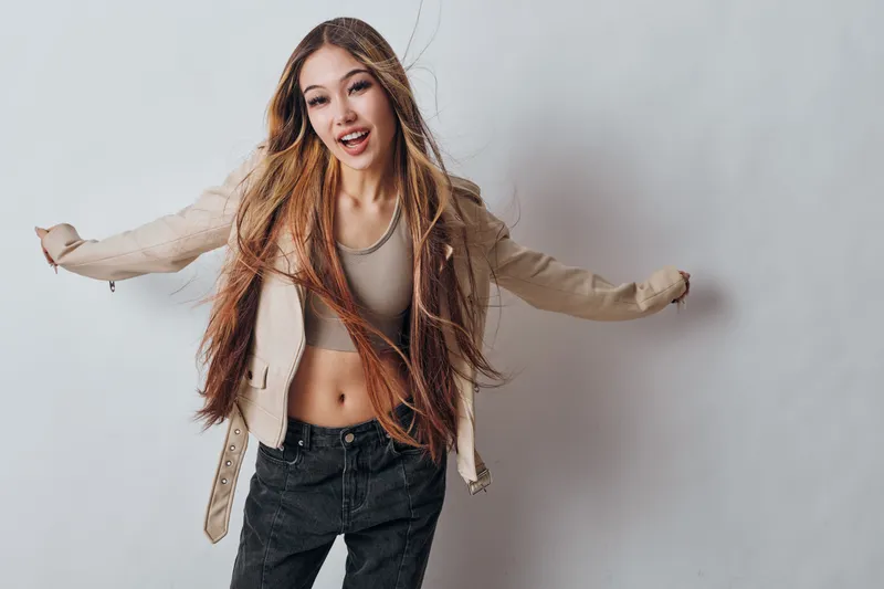 Young woman with long braided hair posing in casual outfit against white background looking happy and confident with stylish jacket and crop top for fashion portrait.