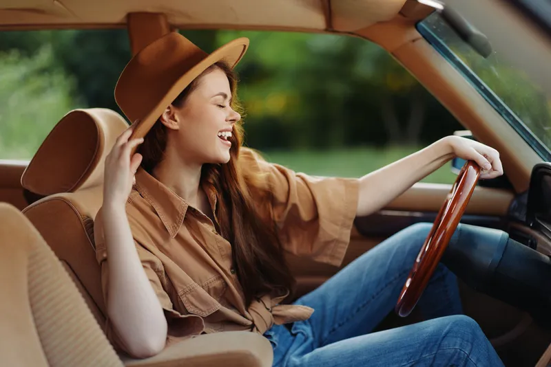 Young woman enjoying a carefree drive in a vintage car, wearing a brown hat, smiles joyfully as she drives in a lush green setting, highlighting a sense of freedom and adventure.