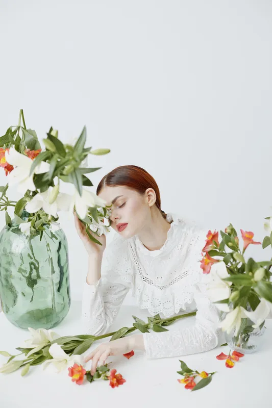 Elegant young woman in white lace dress, surrounded by flowers, expressing serenity and beauty in a soft, bright setting with pastel colors