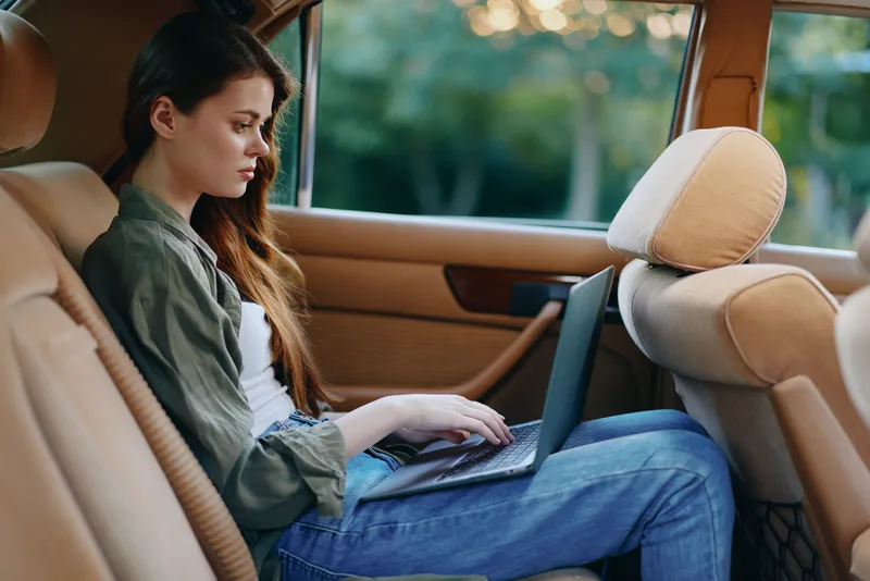 Young woman working on laptop in car during sunset, dressed casually in jeans, reflecting concentration and productivity in a serene outdoor background.