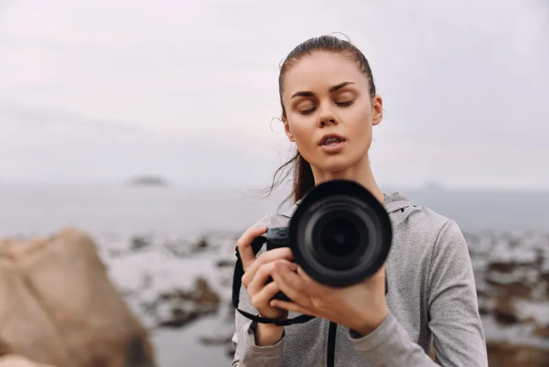 Young woman photographer adjusting camera on the beach, capturing moments Aesthetic mood with cloudy sky and ocean backdrop, showcasing passion for photography