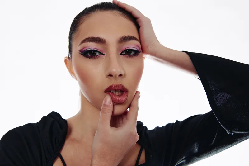 stylish woman with bold makeup in glossy dark lipstick and pastel eyeshadow poses with her hand on her face in a high fashion beauty creative concept photo