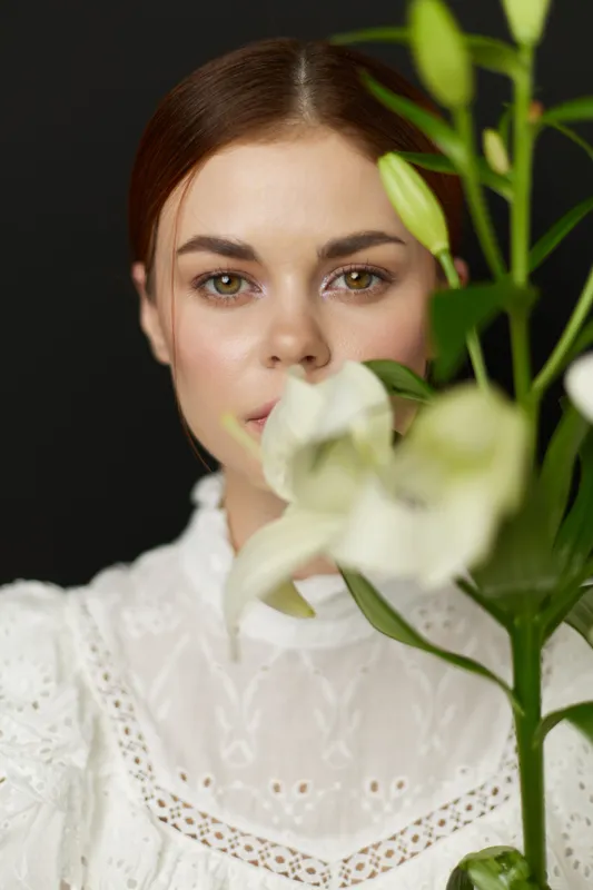 Elegant portrait of a young woman with green eyes holding white lilies against a dark background, showcasing a serene expression and delicate fashion style