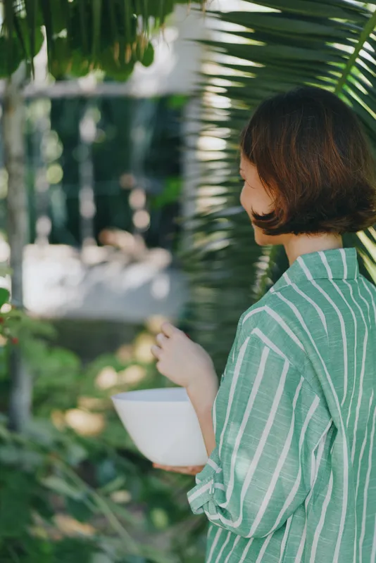 Person wearing a green striped shirt stands among lush greenery, holding a white cup, captured in soft daylight.