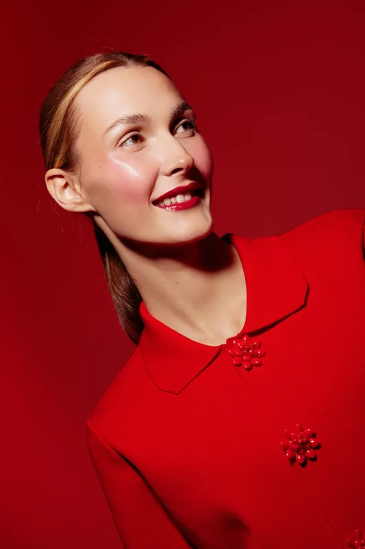 Smiling woman in red coat looks away on red background. Studio portrait with bright makeup and glossy skin. Concept of fashion, beauty, and elegance.
