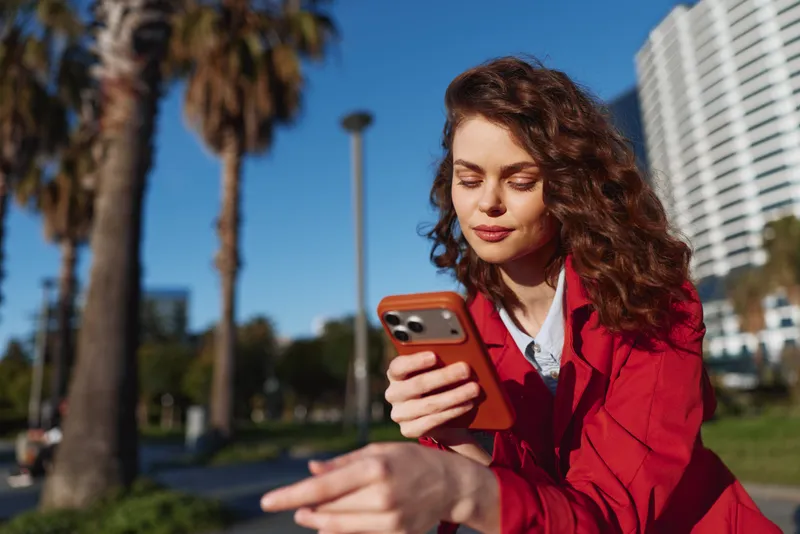 Young woman in red jacket uses smartphone outdoors in sunny park with palm trees and modern buildings in background, focused on screen during daytime.