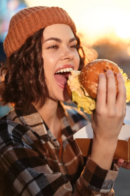 Woman eating a burger outdoors at golden hour, candid smile and beanie, urban street food moment with authenticity, mindful living vibe and emotional storytelling in lifestyle.