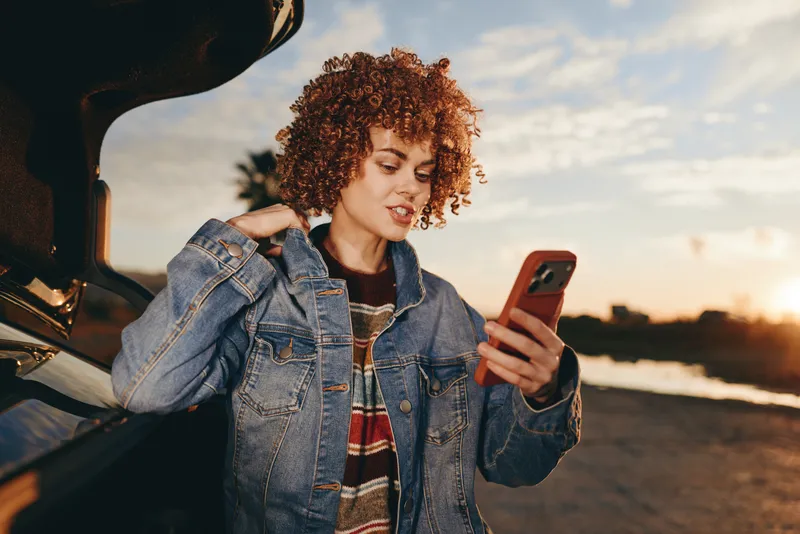 Woman lifestyle outdoors, curly hair, wearing denim jacket, standing by car at sunset, smiling while looking at smartphone, golden hour light, warm sky, relaxed pose