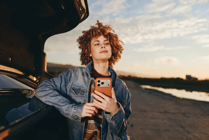 Curly hair woman in denim jacket outdoors at sunset, holding smartphone for selfie beside car, relaxed lifestyle moment, soft warm evening light, cinematic sky, road background