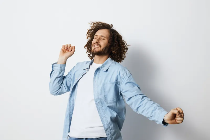 Happy young man dancing joyfully against a light background, wearing a casual denim jacket and a white t shirt, expressing carefree emotions