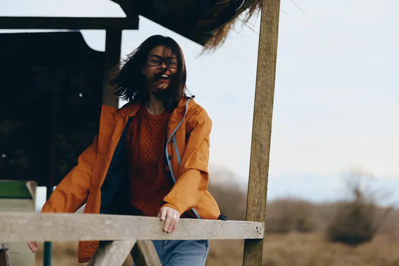 Woman outdoors lifestyle, orange jacket, leaning on wooden fence, smiling with relaxed posture, sunlit rural background, portrait in morning light, soft wind, casual daily moment, warm colors