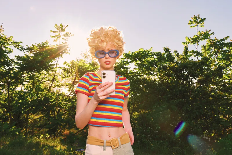 A stylish woman stands outdoors in bright sunlight, wearing a striped crop top and sunglasses, browsing her phone. A carefree lifestyle moment captured with retro film color and bold vibe.