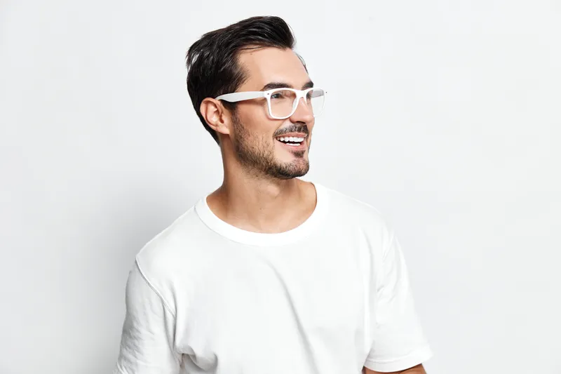 Happy smiling young man with stylish glasses wearing a white t-shirt, looking to the side against a plain solid background in a cheerful studio portrait.