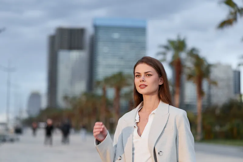 confident woman in light gray coat walking in urban business district with modern skyscrapers and palm trees in background
