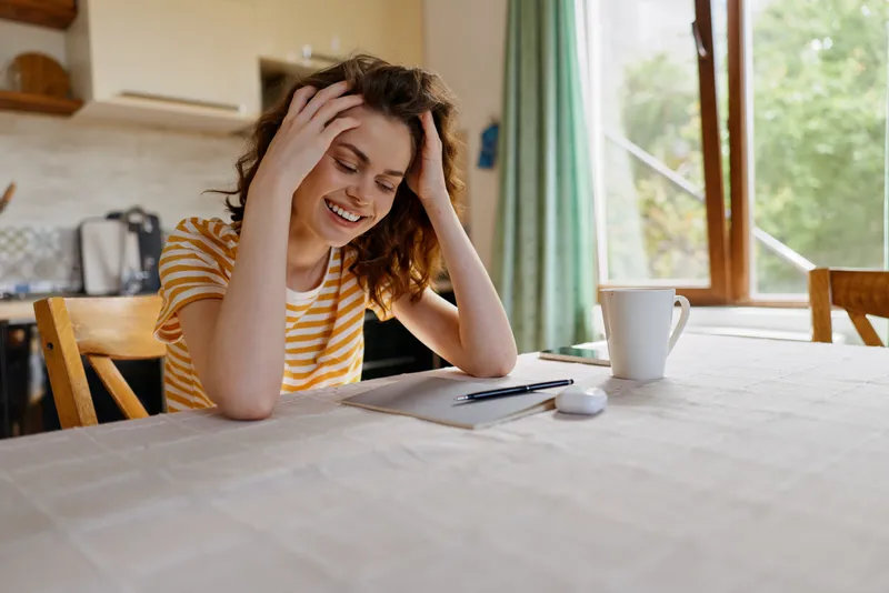 Cheerful woman indoors at home, relaxed lifestyle moment, seated at a table near a bright window, striped shirt, soft natural light, casual mood, everyday tasks