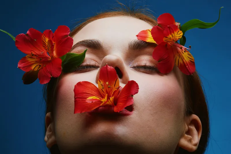 A serene beauty portrait featuring a model with closed eyes surrounded by vibrant red flowers, symbolizing tranquility and nature in a bold blue background