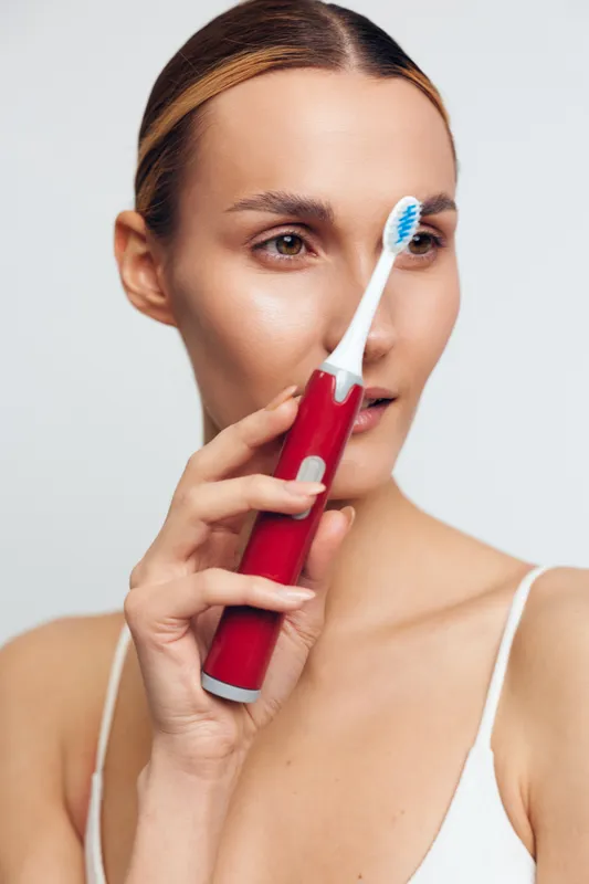 Young woman holding a red electric toothbrush near her face with a white background. Concept of dental hygiene, personal care, and daily routine. Soft natural lighting highlights skin texture.
