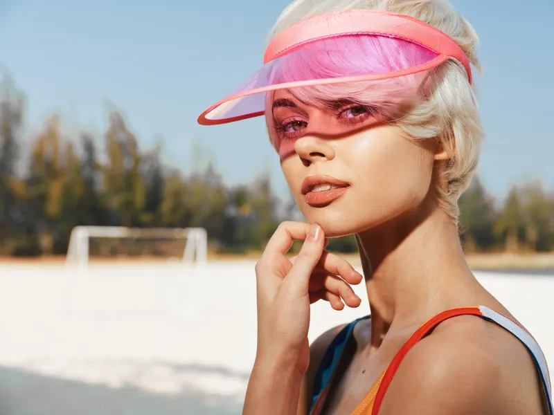 Portrait of a young woman wearing a pink mesh visor on a sunny beach, sunlit skin, relaxed pose, and a soft gaze, capturing a vibrant summer mood.