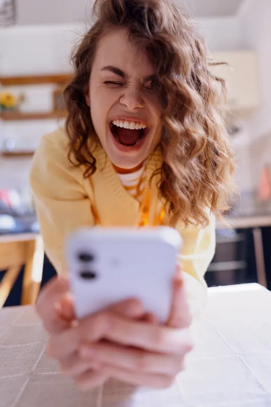 A joyful woman in a casual home setting smiles at a smartphone, capturing a candid selfie. A bright kitchen in the background creates a warm, relaxed lifestyle scene.