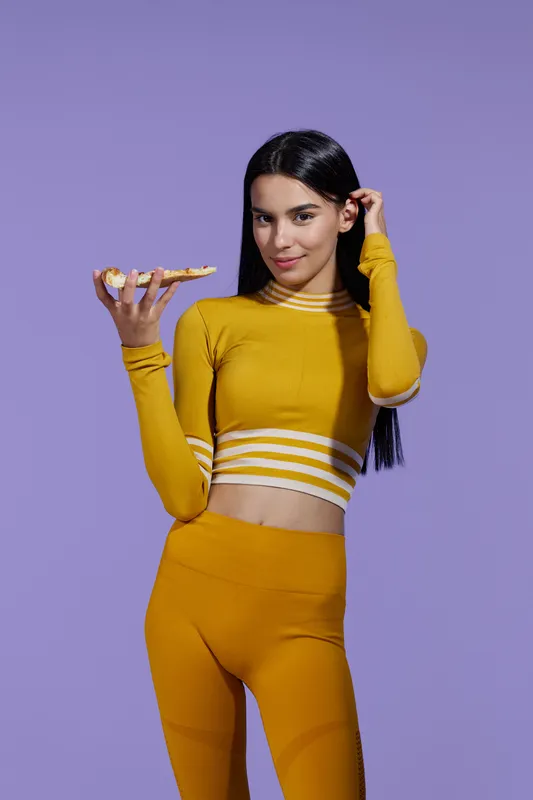 sport fitness woman in yellow activewear holding pizza slice, slim young female with long black hair posing on purple background, healthy lifestyle contrast
