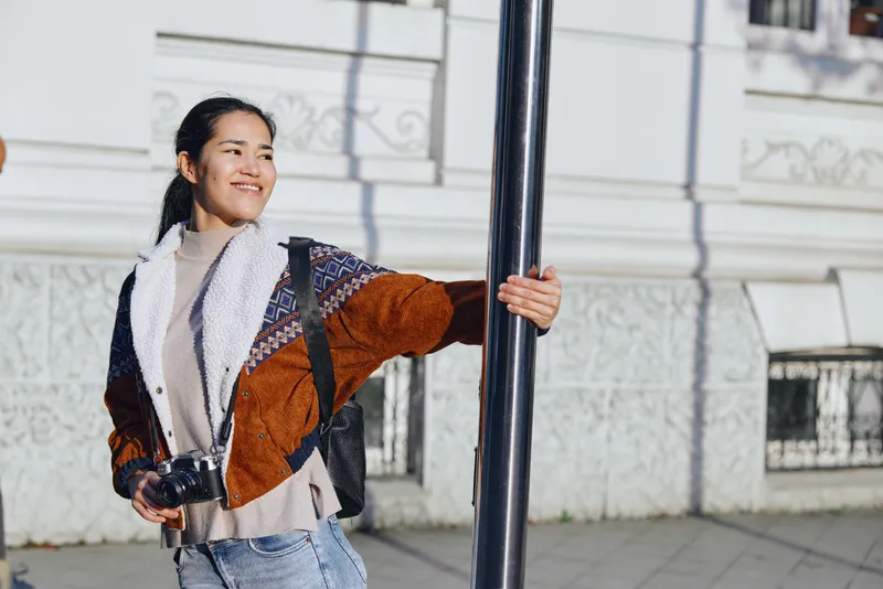 Photographer, woman smiles while holding a camera beside a street pole in a sunny urban setting, casual jacket and denim, candid moment of street photography scene