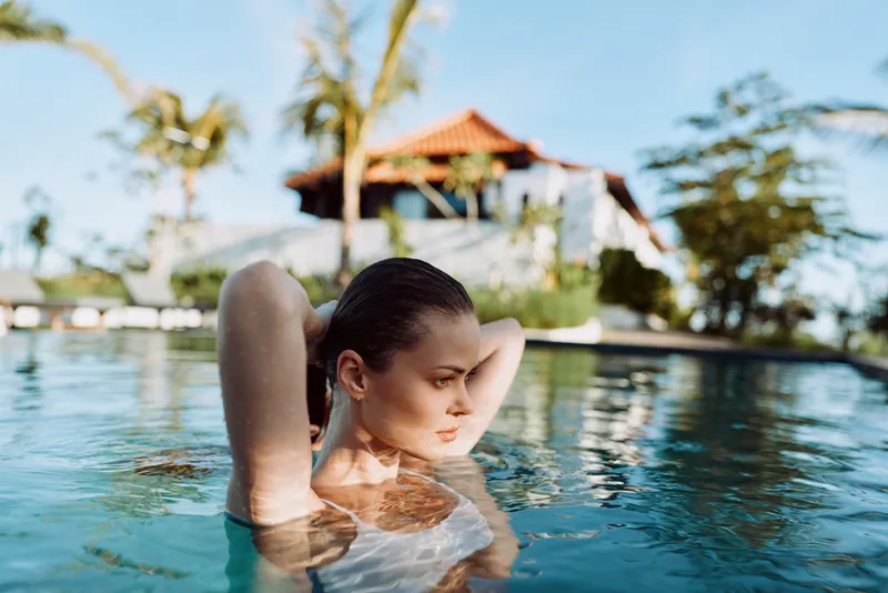 Cheerful woman relaxing in a sunlit pool, arms behind head, relaxed posture, water ripples, warm breeze, tropical backdrop, bright colors, vacation mood, outdoor scene, lifestyle photography