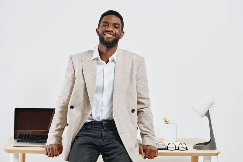 Professional young man with dark skin smiling confidently in office setting wearing beige blazer and white shirt, casual style, modern workspace, natural light, positive work environment, professional