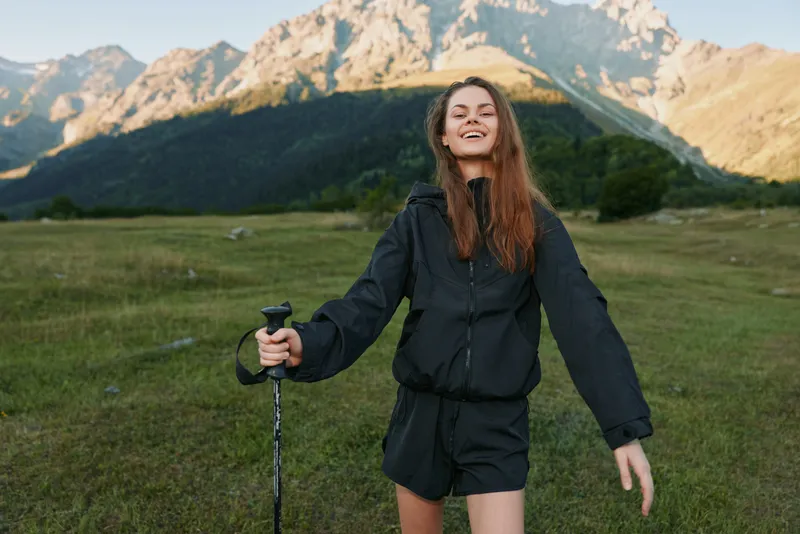 A cheerful hiker smiles broadly as she stands in a wide meadow with distant mountains, holding a trekking pole and enjoying a sunny outdoor adventure.