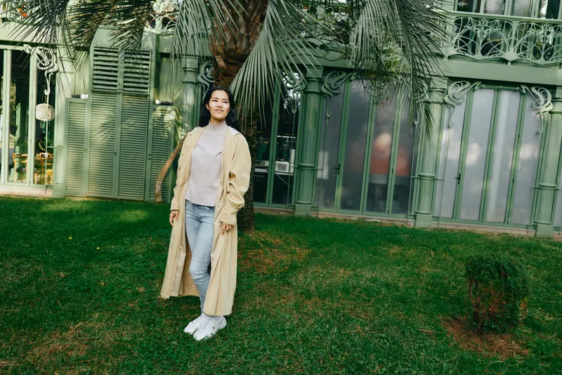 Woman in beige trench coat standing on a green lawn outdoors in a casual fashion scene, relaxed pose near a fence with garden and greenery backdrop, sunlight filtering through trees