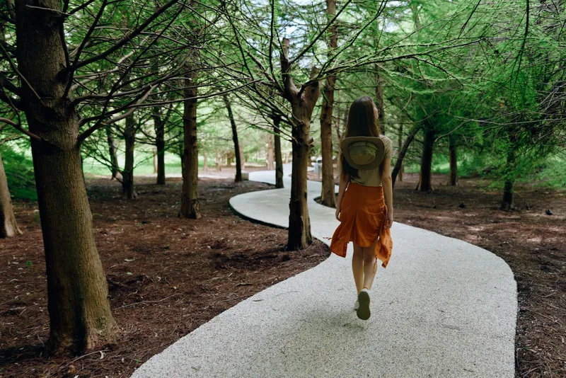 Woman walking on a winding white path through a dense forest with tall trees, wearing orange skirt and backpack. Solitary nature trail, peaceful outdoor journey and exploration.